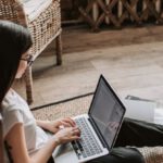 Referral Programs - Young barefoot woman using laptop on floor near books in stylish living room