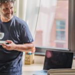 Lean Start-up - Happy adult bearded man in home outfit leaning on table with modern laptop and drinking coffee during break in freelance work on sunny day