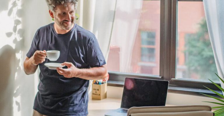 Lean Start-up - Happy adult bearded man in home outfit leaning on table with modern laptop and drinking coffee during break in freelance work on sunny day